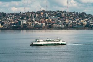 A Washington State Ferry sails on the calm water with Seattle skyline in the background.